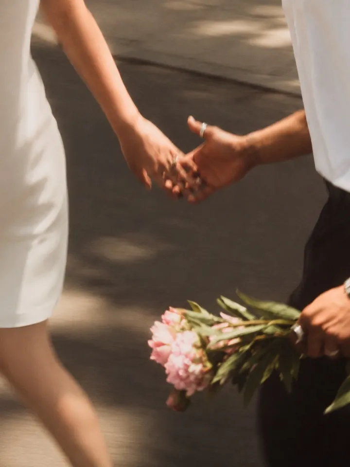 A couple holding hands on a sunlit street, one carrying a bouquet of pink peonies
