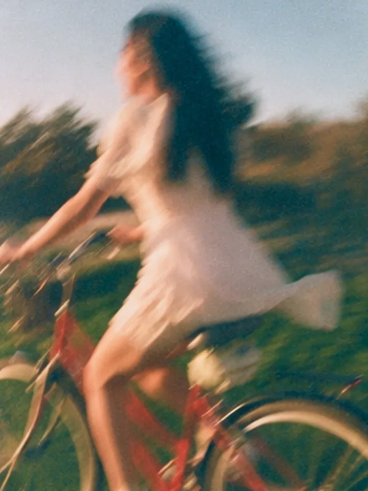 A woman in a white dress riding a bicycle through a sunlit summer field