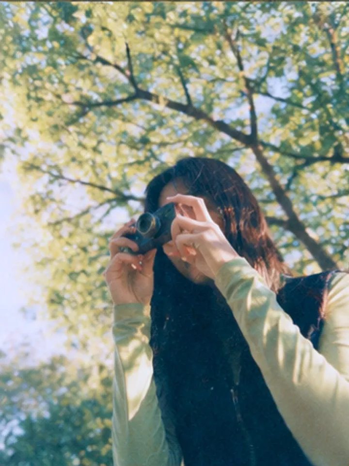 A woman taking a photo with a film camera beneath sunlit trees